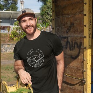 K&ouml;nnte beinhalten: Ein Mann tr&auml;gt ein schwarzes T-Shirt mit einem wei&szlig;en Motiv einer Welle und eines Mondes. Das Motiv befindet sich in einem Kreis mit Sternen drumherum.