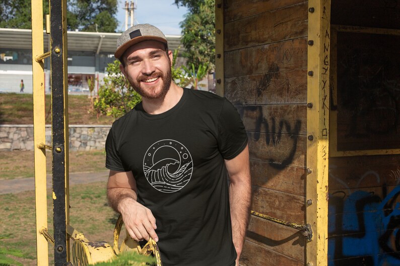 K&ouml;nnte beinhalten: Ein Mann tr&auml;gt ein schwarzes T-Shirt mit einem wei&szlig;en Motiv einer Welle und eines Mondes. Das Motiv befindet sich in einem Kreis mit Sternen drumherum.