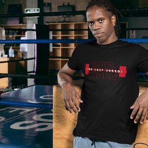 May include: Black t-shirt with the text "BAR WARS The Last Shredi" in red, featuring a barbell graphic. The person in the image is leaning against a wooden surface in a boxing ring.