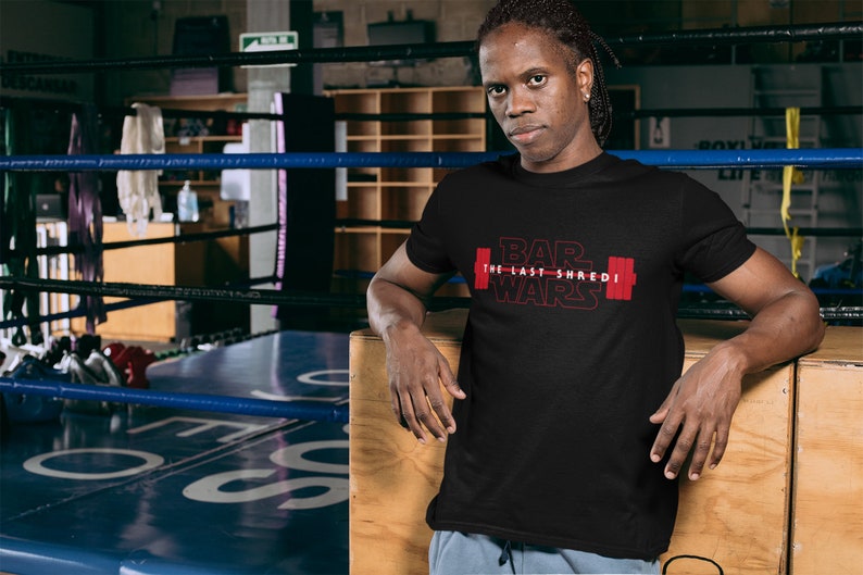 May include: Black t-shirt with the text "BAR WARS The Last Shredi" in red, featuring a barbell graphic. The person in the image is leaning against a wooden surface in a boxing ring.