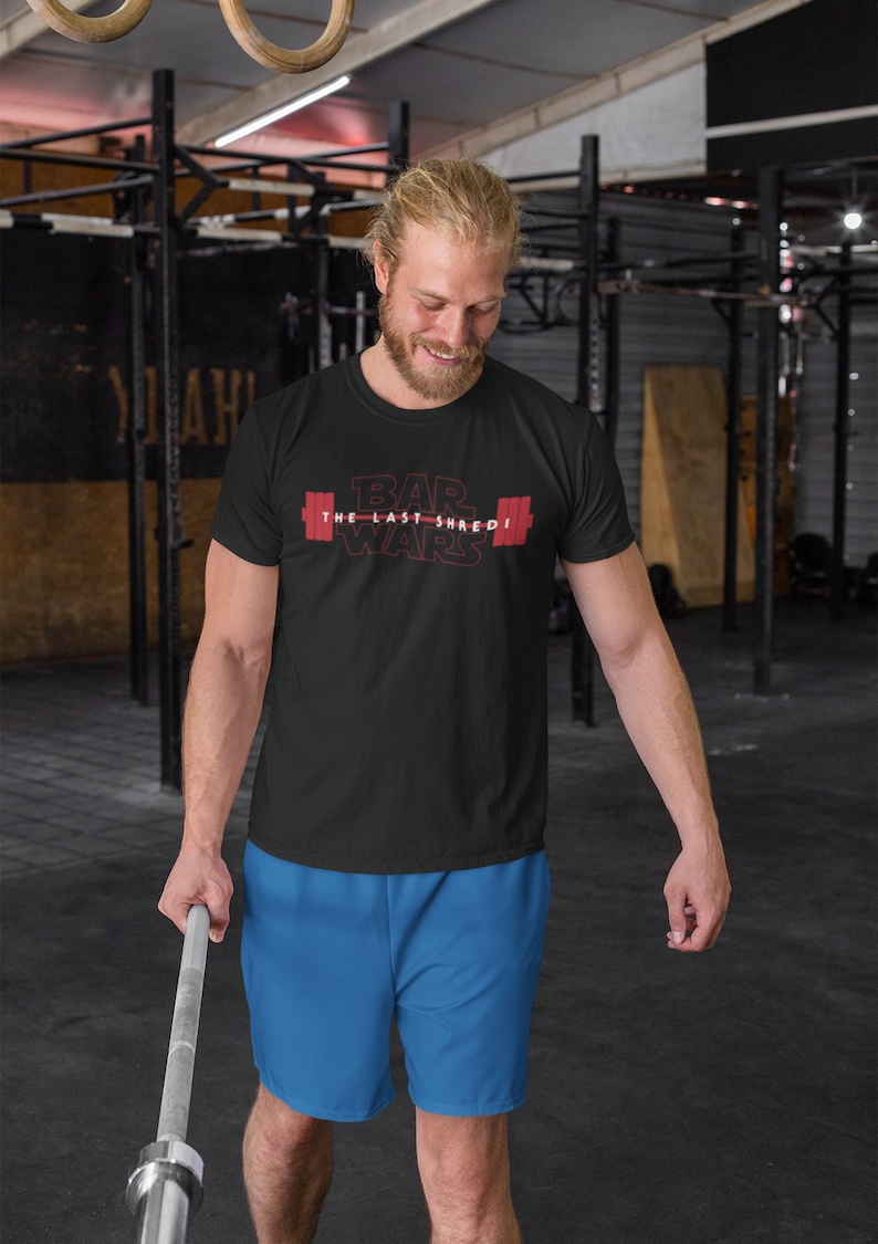 May include: Black t-shirt with the text "BAR WARS The Last Shred" in red, featuring a barbell graphic. The person in the image is wearing blue shorts and holding a barbell in a gym setting.