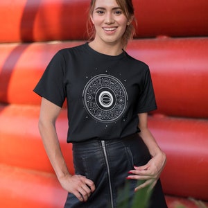 May include: A black t-shirt with a white graphic of a circle with various symbols inside. The graphic is centered on the shirt. The person wearing the shirt is standing in front of a red wall.