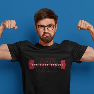 May include: Black t-shirt with the text "BAR WARS THE LAST SHREDI" in red, featuring a barbell graphic. The person in the image is flexing their biceps against a blue backdrop.