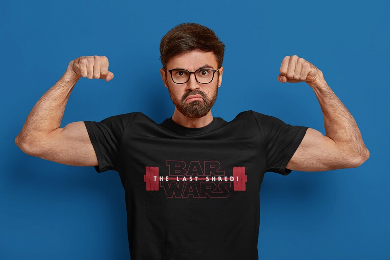 May include: Black t-shirt with the text "BAR WARS THE LAST SHREDI" in red, featuring a barbell graphic. The person in the image is flexing their biceps against a blue backdrop.