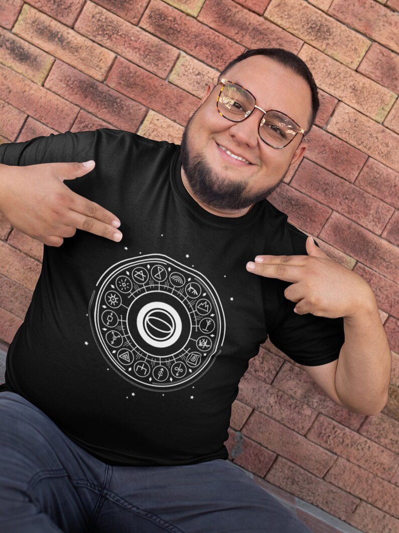 May include: A black t-shirt with a white graphic design of a circle with various symbols inside. The circle is surrounded by stars. The person wearing the shirt is smiling and pointing at the graphic.