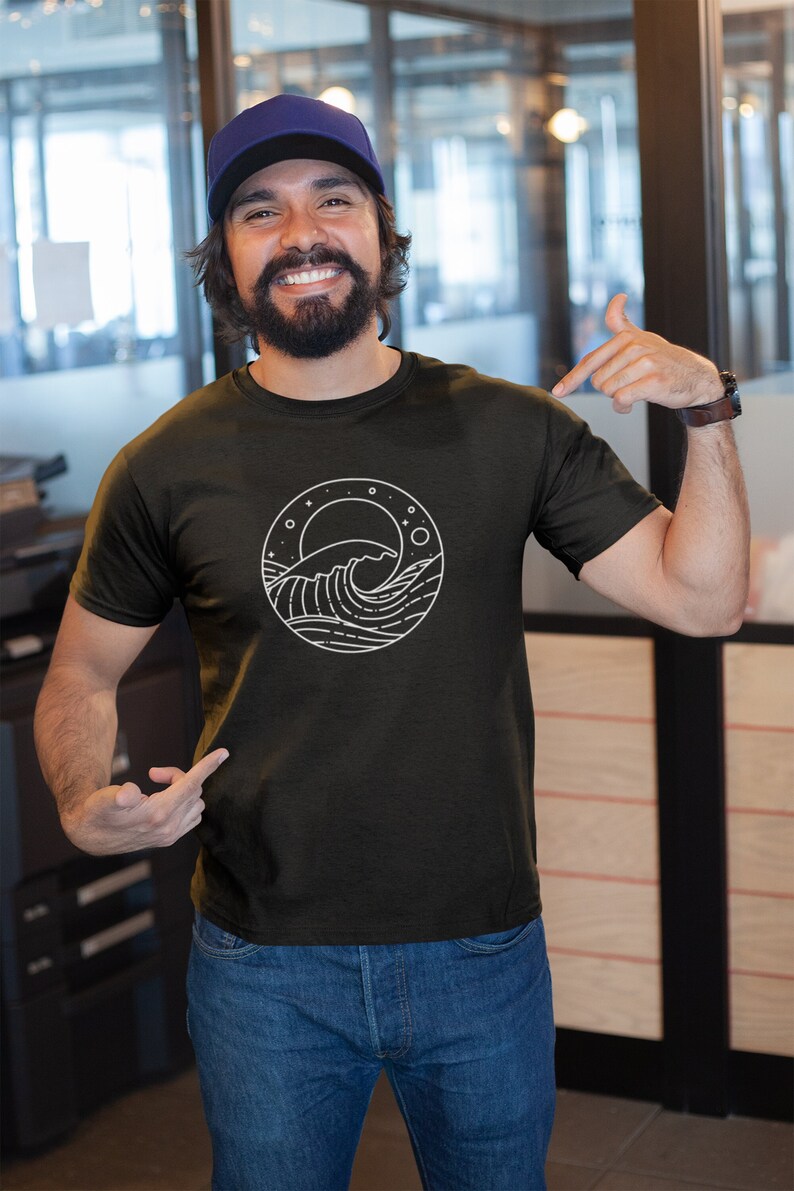 K&ouml;nnte beinhalten: Ein Mann tr&auml;gt ein schwarzes T-Shirt mit einem wei&szlig;en Grafikdruck einer Welle und Sternen in einem Kreis. Er tr&auml;gt eine blaue Baseballkappe und eine blaue Jeans.