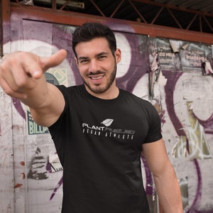 May include: A man wearing a black t-shirt with a white graphic that says "PLANT-POWERED VEGAN ATHLETE". He is standing in front of a graffiti wall and pointing to the camera.