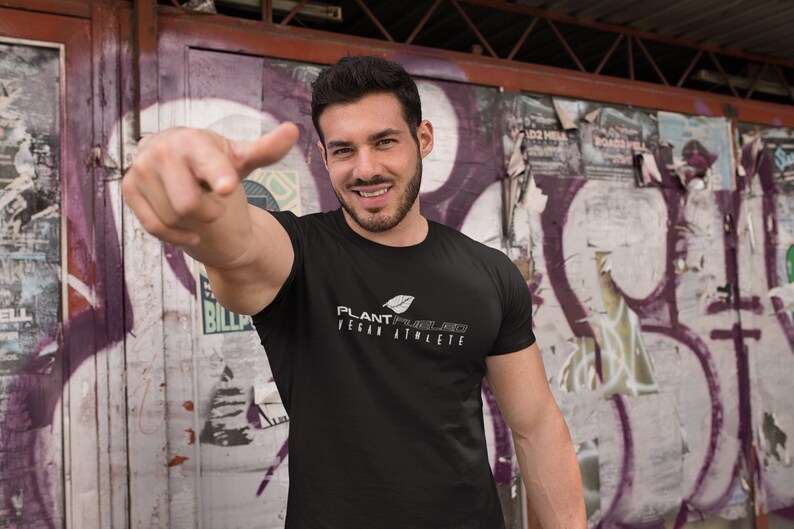 May include: A man wearing a black t-shirt with a white graphic that says "PLANT-POWERED VEGAN ATHLETE". He is standing in front of a graffiti wall and pointing to the camera.