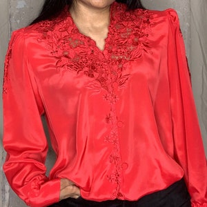Może przedstawiać: Czerwona bluzka z satyny z haftem przypominającym koronkę wokół kołnierzyka i z przodu. Bluzka ma długie rękawy i zapięcie na guziki z przodu.