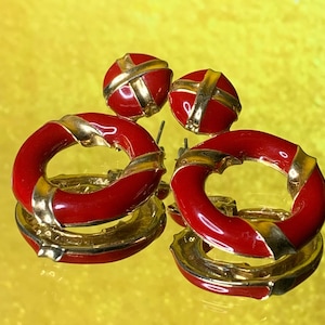 May include: A set of vintage earrings with a red enamel finish and gold-tone detailing. The set includes hoop earrings and stud earrings. The hoop earrings have a circular design, and the studs are round. The earrings are displayed on a reflective surface, reflecting the light.