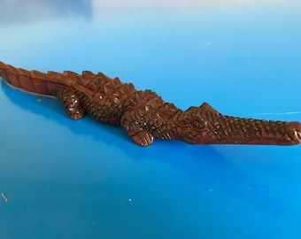 May include: A detailed, wooden alligator figurine, carved in a rich, reddish-brown hue. The reptile is depicted in a realistic pose, with textured scales and a long snout. The figurine is a decorative item.