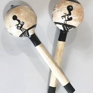 May include: Two wooden maracas with a light brown finish and black painted details. The round heads feature a black and white design, and the handles are crossed. The handles are light-colored wood with black tips.