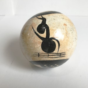 May include: A hand-painted, spherical decorative stone. The stone features a beige background with black abstract art. The artwork includes a musical note-like design and geometric shapes. The stone is smooth and appears to be varnished.
