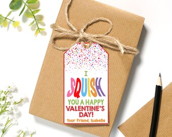 Pode incluir: Uma etiqueta de presente colorida para o Dia dos Namorados com fundo branco e confete. A etiqueta diz "I SQUISH YOU A HAPPY VALENTINE'S DAY!" e "Your Friend, Isabella".