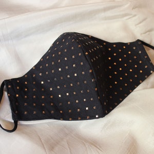May include: Black face mask with a pattern of small, gold-colored squares. The mask has adjustable black ear loops and is made of a soft, flexible material. The mask is designed to cover the nose and mouth.