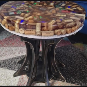 Puede incluir: Mesa de centro redonda con una tapa de resina transparente llena de corchos de vino. La mesa tiene una base de metal oscuro con un diseño curvo único. La tapa de la mesa tiene aproximadamente 91 cm de diámetro.