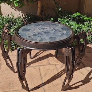 Könnte beinhalten: Ein runder Beistelltisch aus dunklem Metall mit einer gemusterten Glasplatte. Der Tisch hat ein einzigartiges Design mit sechs dekorativen Beinen. Der Tisch hat einen Durchmesser von ca. 61 cm und ist 51 cm hoch.