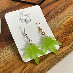 May include: A pair of lime green leaf-shaped earrings with silver and gold-coloured hooks. The earrings are displayed on a white card with the text "POURED By Addy" and a floral design. The background is a wooden surface.