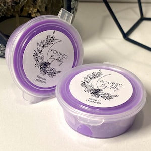 May include: Two small, round, lavender-colored wax melt containers with clear lids. Each container has a white label with the text "POURED By Addy" and "FRENCH LAVENDER" in black. The labels also feature a floral and crescent moon design.