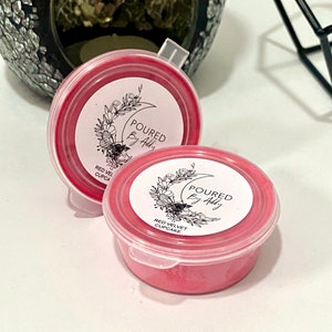 May include: Two small, round, red wax melt containers with clear lids. Each container has a white label with the text "Poured By Addy" and "Red Velvet Cupcake" in black. The background includes a decorative candle holder.