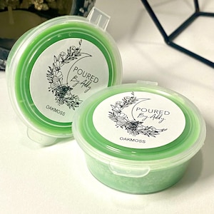 May include: Two small, round, green wax melt containers with clear lids. Each container has a white label with the text "POURED By Addy" and "OAKMOSS" with a floral crescent moon design. The wax melts are a light green color.
