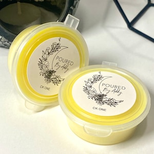 May include: Two round, yellow wax melt containers with clear lids. Each container has a white label with floral and crescent moon graphics and the text "POURED By Addy" and "CK ONE". The containers are on a white surface.
