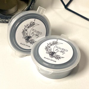 May include: Two small, round, black wax melt containers with clear lids. Each container has a white label with the text "POURED By Addy" and "Black Opium" printed on it, along with a floral design. The containers are on a white surface.