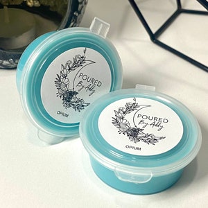 May include: Two round, light blue wax melt containers with clear lids. Each container has a white label with the text "POURED By Addy" and "OPIUM" below a floral and crescent moon design. The containers are on a white surface.