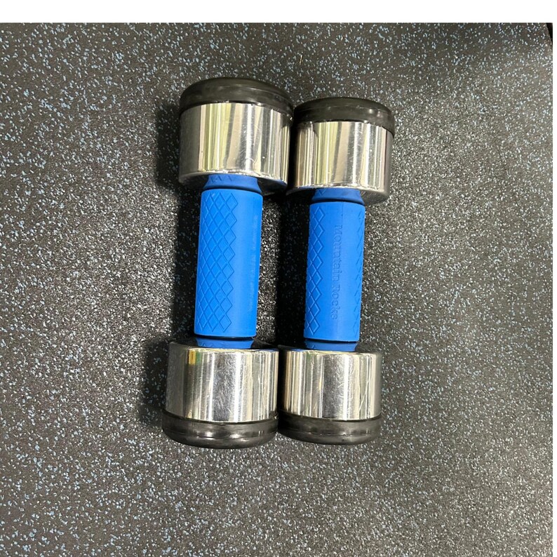 Puede incluir: Un par de mancuernas plateadas con mangos de goma azul. Las mancuernas est&aacute;n sobre una superficie negra texturizada.