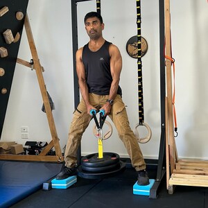 Puede incluir: Un hombre con un tanque negro y pantalones caqui est&aacute; usando una m&aacute;quina de cable para levantar pesas. Est&aacute; de pie sobre dos plataformas azules y blancas. La m&aacute;quina de cable est&aacute; conectada a una pila de pesas con placas negras.