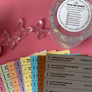 Op de afbeelding: Een doorzichtige dankbaarheids pot met een label met de tekst "Pick Me When". De pot is omgeven door kleurrijke kaarten met inspirerende citaten en doorzichtige vlinderdecoraties. De pot staat op een roze ondergrond.
