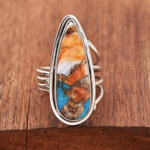May include: A silver ring with a teardrop-shaped stone. The stone is a mix of orange, white, and blue colors.