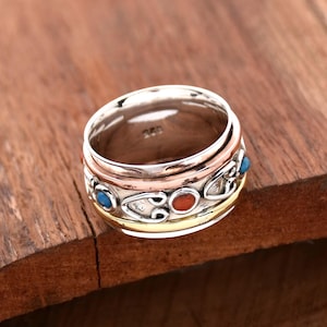 Może przedstawiać: Obrócający się pierścień ze srebra, miedzi i złota z niebieskimi i czerwonymi kamieniami. Pierścień ma ozdobny wzór z sercami i spiralami.