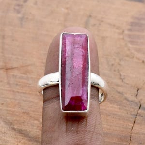 Op de afbeelding: Een zilveren ring met een grote, rechthoekige, robijnkleurige edelsteen. De edelsteen is gezet in een gepolijste zilveren band. Het ontwerp van de ring is eenvoudig en benadrukt de levendige kleur en vorm van de steen.
