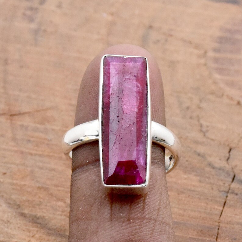 Op de afbeelding: Een zilveren ring met een grote, rechthoekige, robijnkleurige edelsteen. De edelsteen is gezet in een gepolijste zilveren band. Het ontwerp van de ring is eenvoudig en benadrukt de levendige kleur en vorm van de steen.