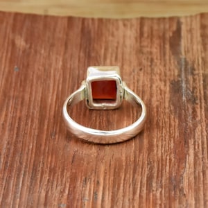 Pode incluir: Um anel de prata com uma pedra preciosa quadrada, vermelho-alaranjado. A banda do anel &eacute; lisa e arredondada, e a pedra preciosa &eacute; engastada numa moldura quadrada. O anel &eacute; exibido numa superf&iacute;cie de madeira.