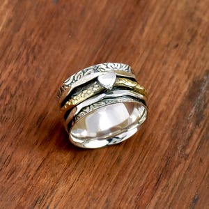 Peut inclure: Une bague spinner en argent avec un accent en forme de cœur et des bandes dorées. La bague présente des motifs gravés complexes et une finition polie, sur une surface en bois chaleureux. La bague est conçue pour tourner.