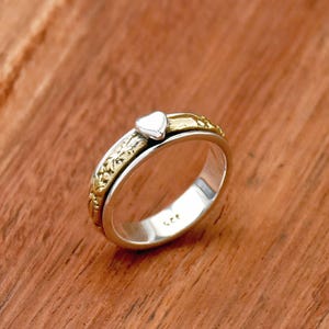 May include: A silver spinning ring with a gold band and a small heart charm. The ring is on a wooden surface.