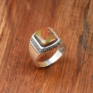 May include: A silver ring featuring a square, multi-coloured stone. The stone displays shades of green, orange, and brown. The ring has a decorative border with a repeating geometric pattern. The ring is set on a wooden surface.