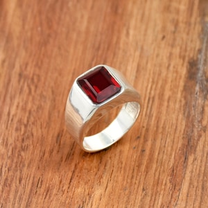 May include: A silver ring with a large, rectangular, deep red gemstone. The ring has a wide band and a polished finish. The gemstone is set in a square bezel, and the ring is displayed on a wooden surface.