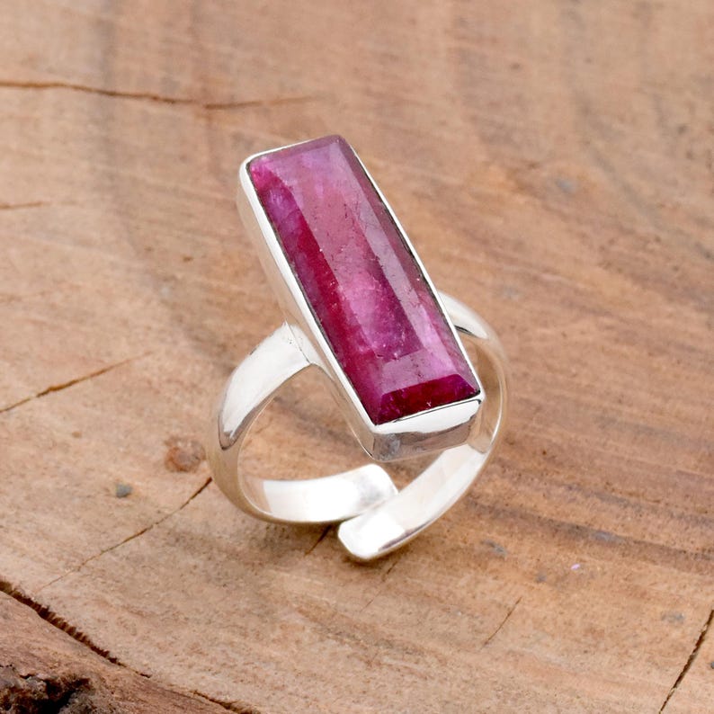 Op de afbeelding: Een zilveren ring met een grote, rechthoekige, dieproze robijn edelsteen. De ring heeft een open, verstelbare band. De robijn is gezet in een bezel setting, die de levendige kleur benadrukt. Gepresenteerd op een houten oppervlak.