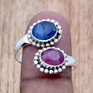 May include: A silver ring with two oval-shaped gemstones. The top stone is a blue sapphire, and the bottom stone is a pink ruby. Both stones are set in a silver bezel with a beaded edge.
