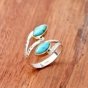 Puede incluir: Un anillo de plata con dos piedras de color turquesa, de talla marquesa, engastadas en biseles dorados. El anillo tiene una textura martillada en la banda y se muestra sobre una superficie de madera.