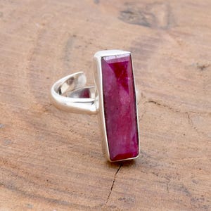 Op de afbeelding: Een zilveren ring met een rechthoekige, dieprode edelsteen. De edelsteen is gezet in een zilveren zetting en de ring heeft een verstelbare band. De ring wordt getoond op een houten oppervlak.