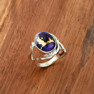 Può includere: Anello in argento con pietra ovale viola con macchie dorate. L'anello presenta un design traforato sui lati e una finitura lucida. L'anello è appoggiato su una superficie di legno.