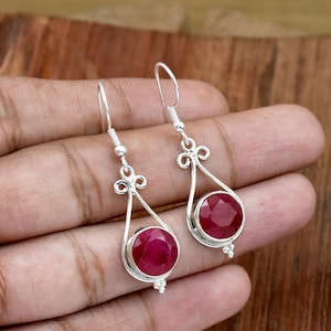 Puede incluir: Un par de pendientes colgantes de plata con grandes piedras preciosas redondas de color rojo intenso. Los pendientes tienen un diseño calado decorativo con forma de lágrima y un pequeño grupo de cuentas.