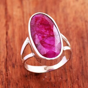 Peut inclure: Bague en argent ornée d'un grand rubis ovale. Le rubis est d'un rose profond avec des facettes visibles. La bague a un design simple et élégant avec une monture fendue.