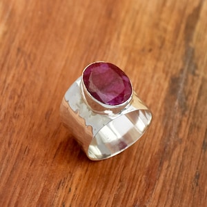 May include: A wide silver ring featuring a large, oval ruby gemstone. The ring has a hammered texture on the band and a polished finish. The ruby is a deep red colour, set in a bezel setting.