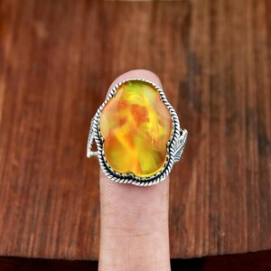 May include: A silver ring featuring a large, oval-shaped gemstone with vibrant yellow and orange hues. The gemstone is set in a detailed silver band with a rope-like border and leaf accents, creating a unique and eye-catching design.