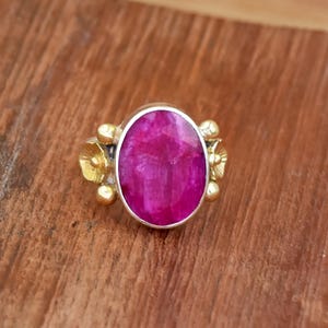 Puede incluir: Un anillo de plata con una gran gema ovalada de color magenta como pieza central. El anillo presenta detalles florales dorados a cada lado de la gema, con pequeñas cuentas doradas. El anillo está sobre un fondo de madera.